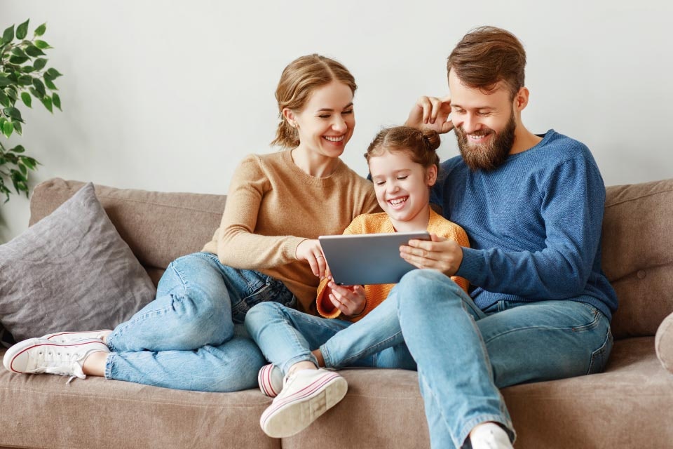 Eine glückliche Familie sitzt auf einem Sofa und schaut gemeinsam auf ein Tablet, während sie lächeln; die Eltern tragen gemütliche Kleidung und sitzen nebeneinander mit ihrem Kind in der Mitte.