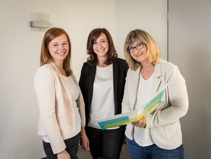 Drei lächelnde Frauen in formeller Kleidung stehen nebeneinander in einem Büro; eine hält eine geöffnete Mappe mit Papieren.