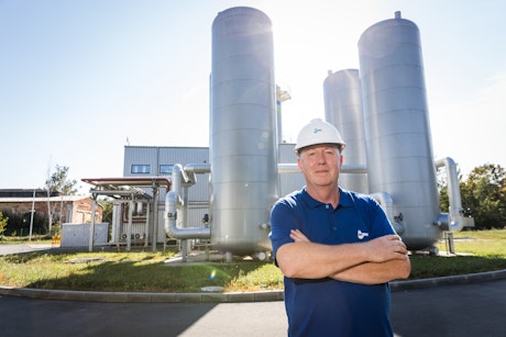 Ein Ingenieur steht mit verschränkten Armen vor großen silbernen Industrieanlagen und einem Gebäude unter blauem Himmel, Helm tragend.