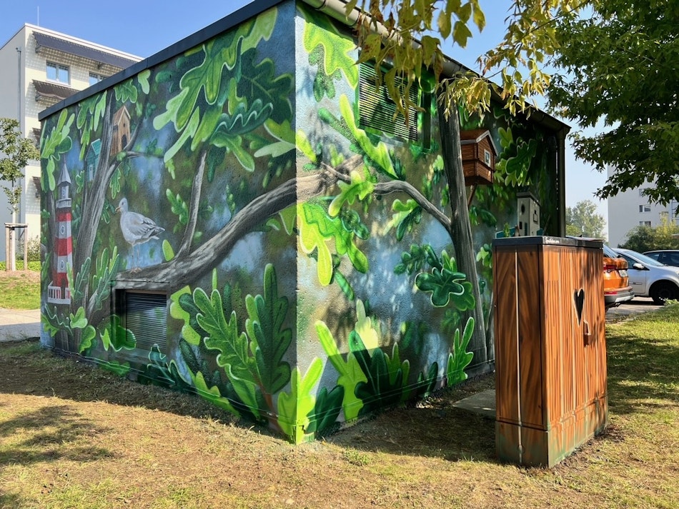Ein kleines Gebäude ist mit einem lebhaften Wandgemälde bemalt, das grüne Blätter, eine Möwe, Vogelhäuser und einen Leuchtturm zeigt, umgeben von einem gepflegten Rasen und einem Baum bei sonnigem Wetter.