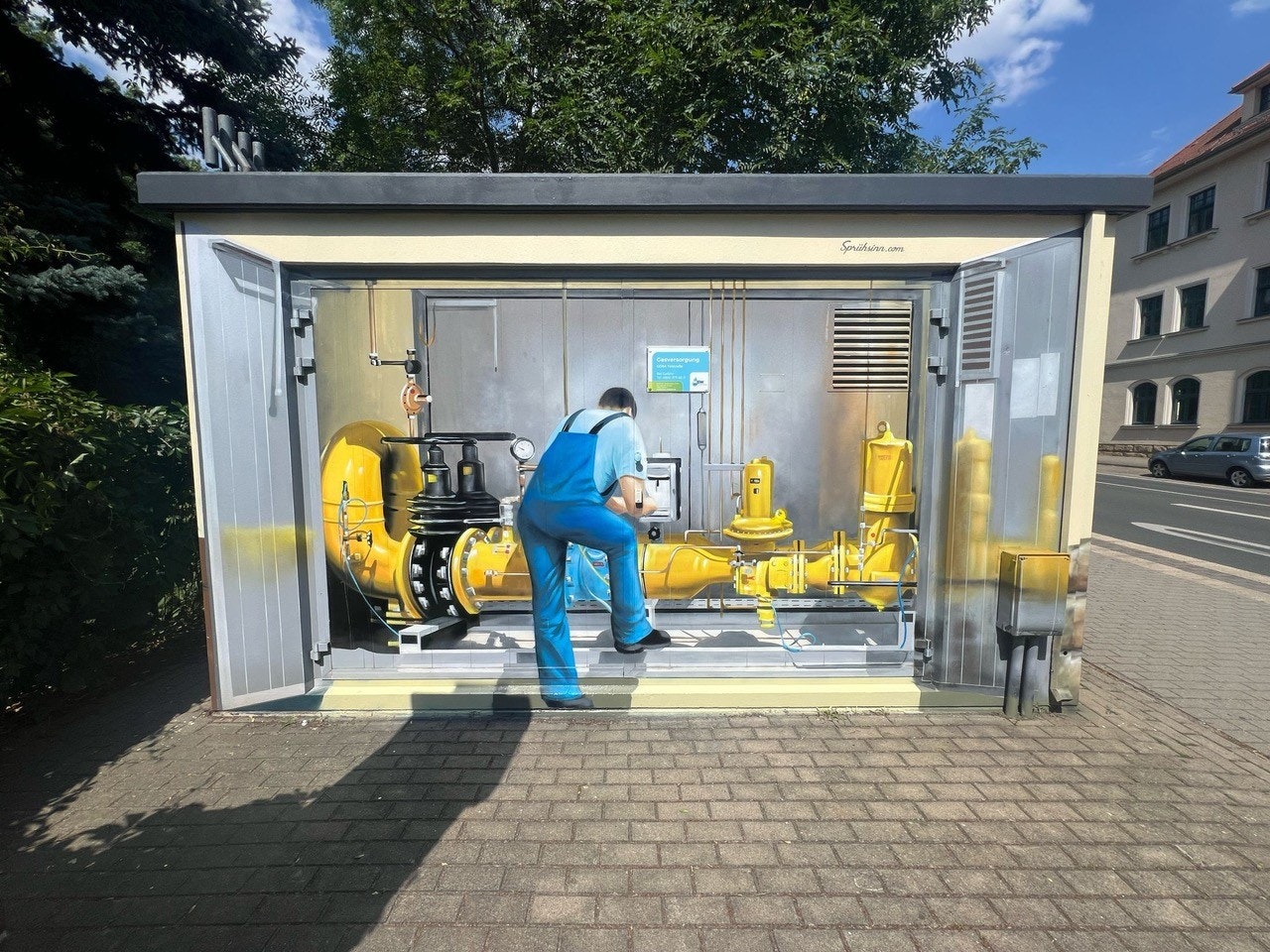 Ein Wandbild auf einer Metalltür zeigt einen Mann in blauer Arbeitskleidung, der mit Maschinen im Inneren eines Technikraums arbeitend dargestellt ist; realistische Details lassen die gemalten Objekte dreidimensional erscheinen.