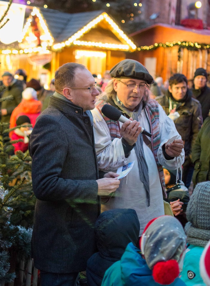 Eine festliche Weihnachtsmarkt-Szene mit Menschen in winterlicher Kleidung, während ein Mann mit Mikrofon eine Ansprache hält, umgeben von hell erleuchteten Holzständen und festlichen Dekorationen.