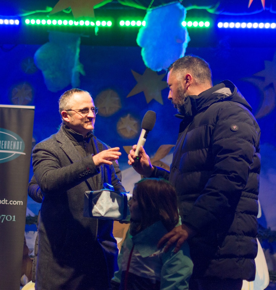 Eine Veranstaltung in einem festlich beleuchteten Raum zeigt einen Mann, der einem anderen ein Mikrofon überreicht; im Hintergrund sind bunte Dekorationen und ein Banner mit einem Unternehmenslogo zu sehen.
