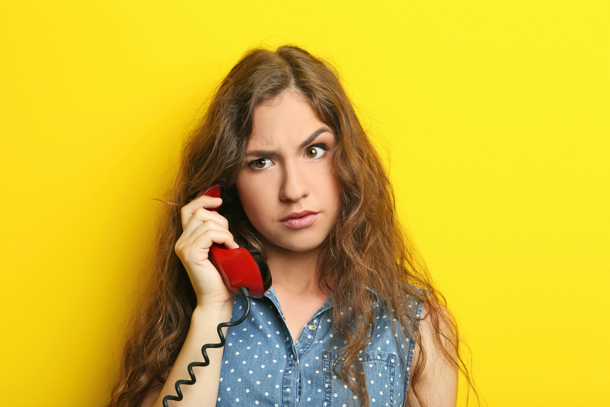 Eine Frau mit langem braunem Haar und einem ärmellosen, blauen Punktekleid hält ein rotes Festnetztelefon an das Ohr, während sie nachdenklich vor einem leuchtend gelben Hintergrund steht.