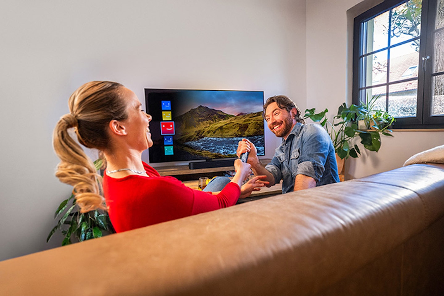 Eine Frau und ein Mann sitzen auf einem braunen Ledersofa in einem Wohnzimmer vor einem eingeschalteten Fernseher, lachend und Händchen haltend, mit Pflanzen und einem beleuchteten Fenster im Hintergrund.
