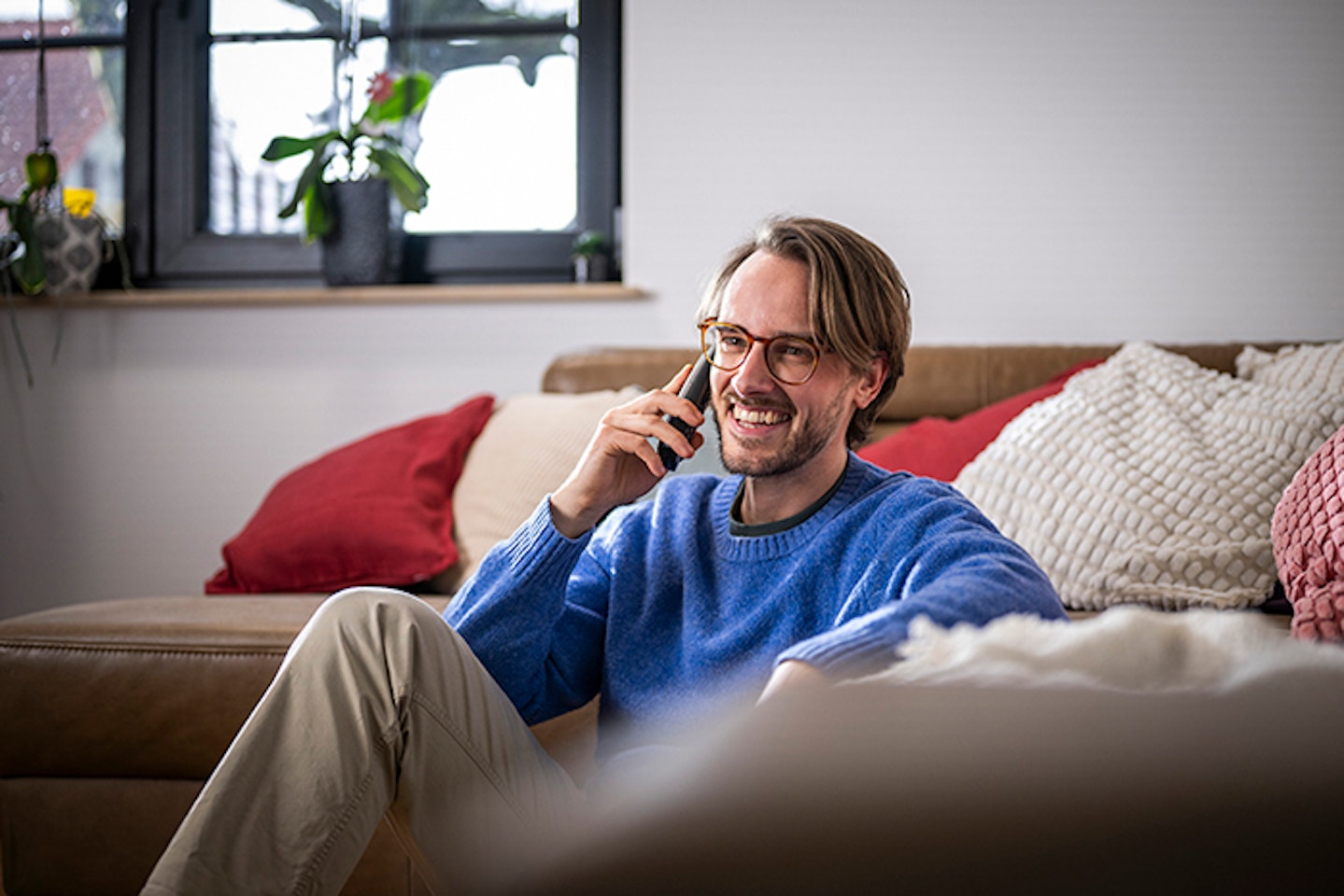 Ein Mann sitzt entspannt auf einem beigen Ledersofa in einem gemütlichen Wohnzimmer und lacht, während er ein Telefongespräch führt; im Hintergrund stehen Pflanzen auf einer Fensterbank.
