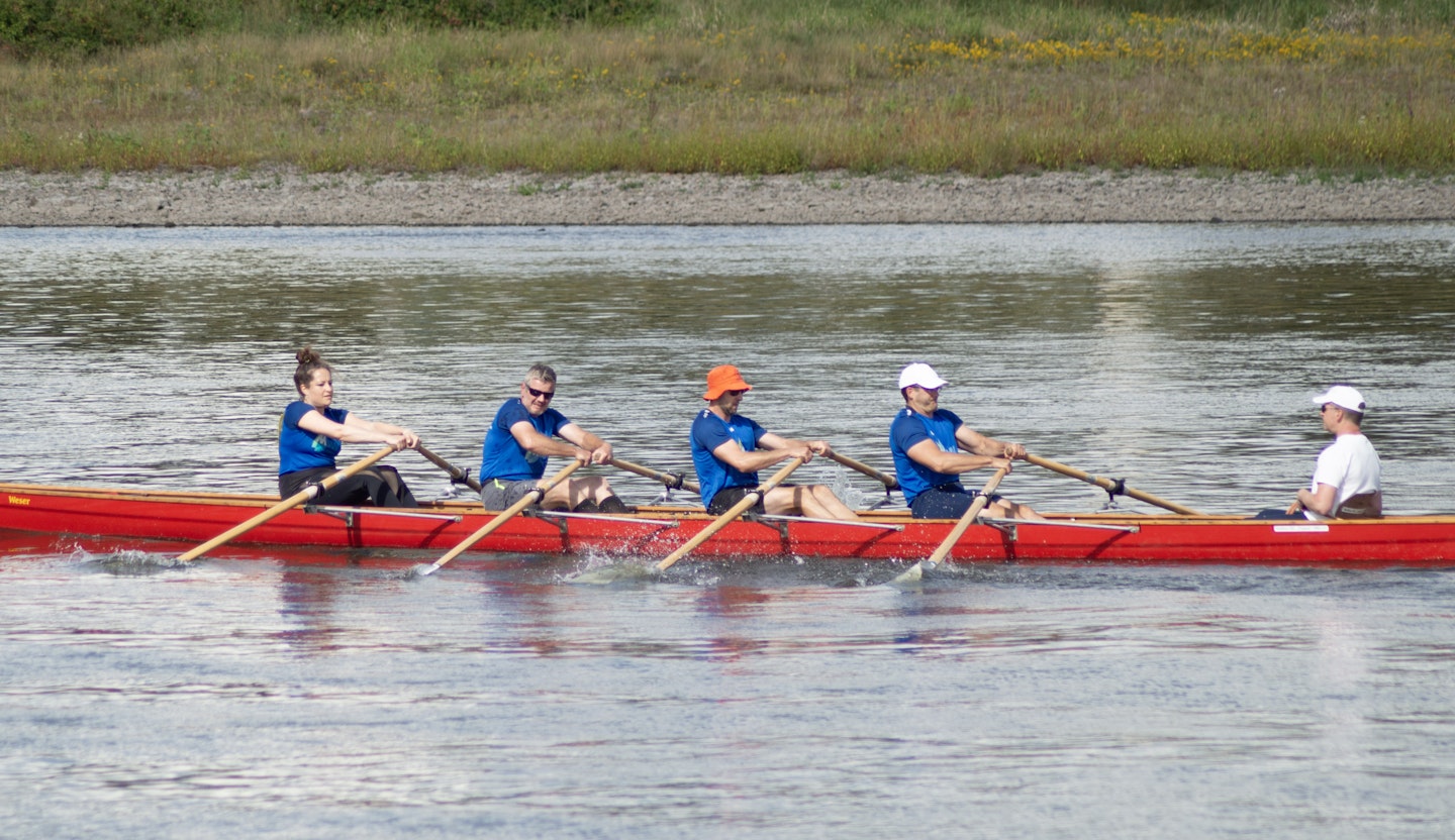 Eine Gruppe von fünf Personen in blauen T-Shirts rudert synchron in einem langen roten Ruderboot auf einem ruhigen Fluss, umgeben von einer natürlichen Uferlandschaft.