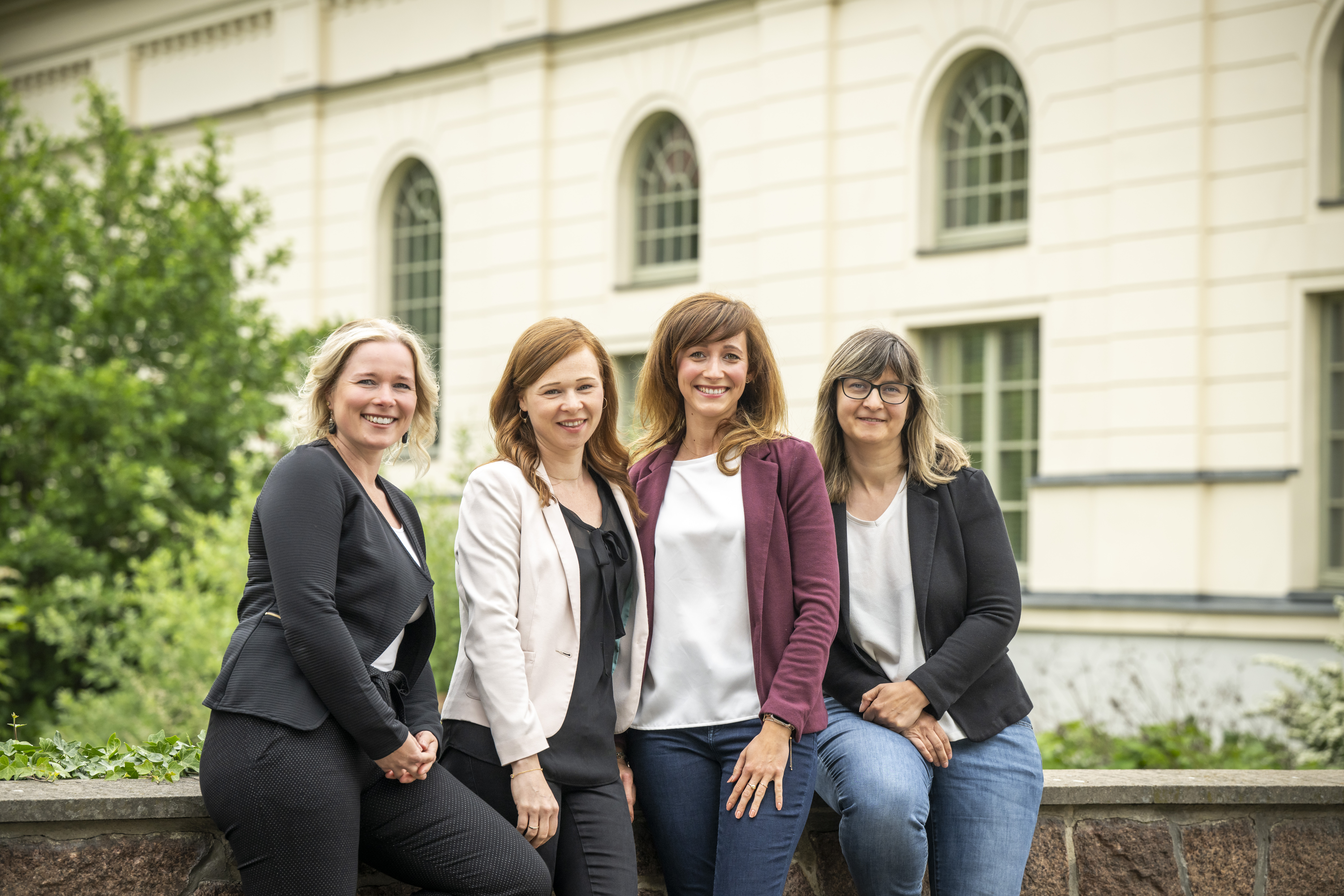 Vier Personen in Businesskleidung stehen lächelnd vor einem historischen Gebäude mit großen Fenstern und einem Garten, während sie auf einer niedrigen Mauer posiert sind.
