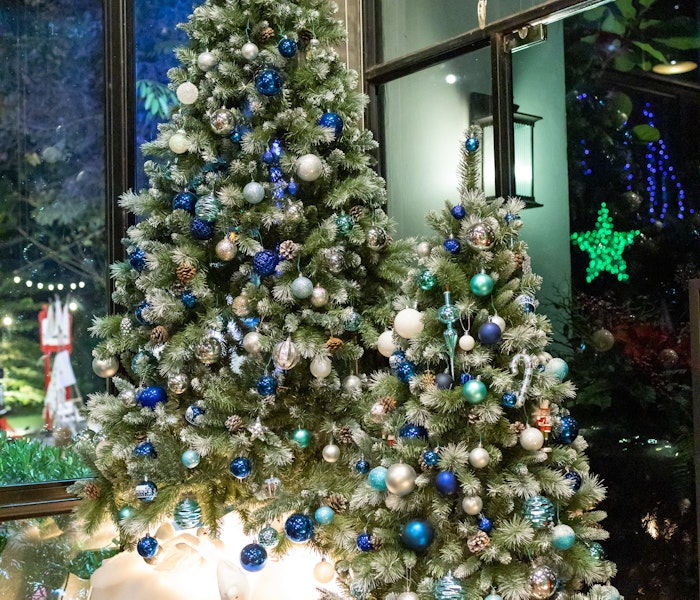 Zwei festlich geschmückte Weihnachtsbäume stehen in einem modernen Raum mit großen Fenstern, die in eine nächtliche, mit Lichtern erleuchtete Landschaft blicken; die Bäume sind mit blauen und silbernen Kugeln dekoriert.