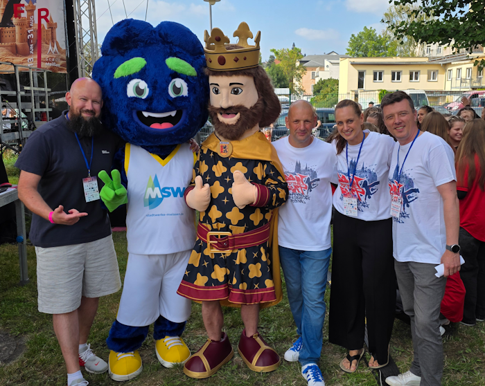 In einem Freiluft-Event stehen zwei Maskottchen – eines in Form einer Krone und eines mit einem blauen Gesicht – zusammen mit einer kleinen Gruppe fröhlicher Menschen vor einem Hintergrund aus Zelte und Bäumen.