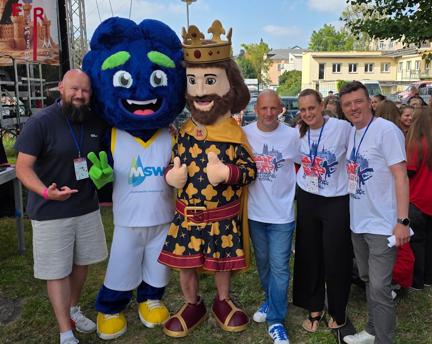 In einem Freiluft-Event stehen zwei Maskottchen – eines in Form einer Krone und eines mit einem blauen Gesicht – zusammen mit einer kleinen Gruppe fröhlicher Menschen vor einem Hintergrund aus Zelte und Bäumen.