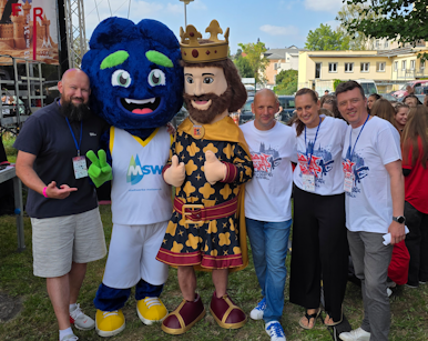 In einem Freiluft-Event stehen zwei Maskottchen – eines in Form einer Krone und eines mit einem blauen Gesicht – zusammen mit einer kleinen Gruppe fröhlicher Menschen vor einem Hintergrund aus Zelte und Bäumen.