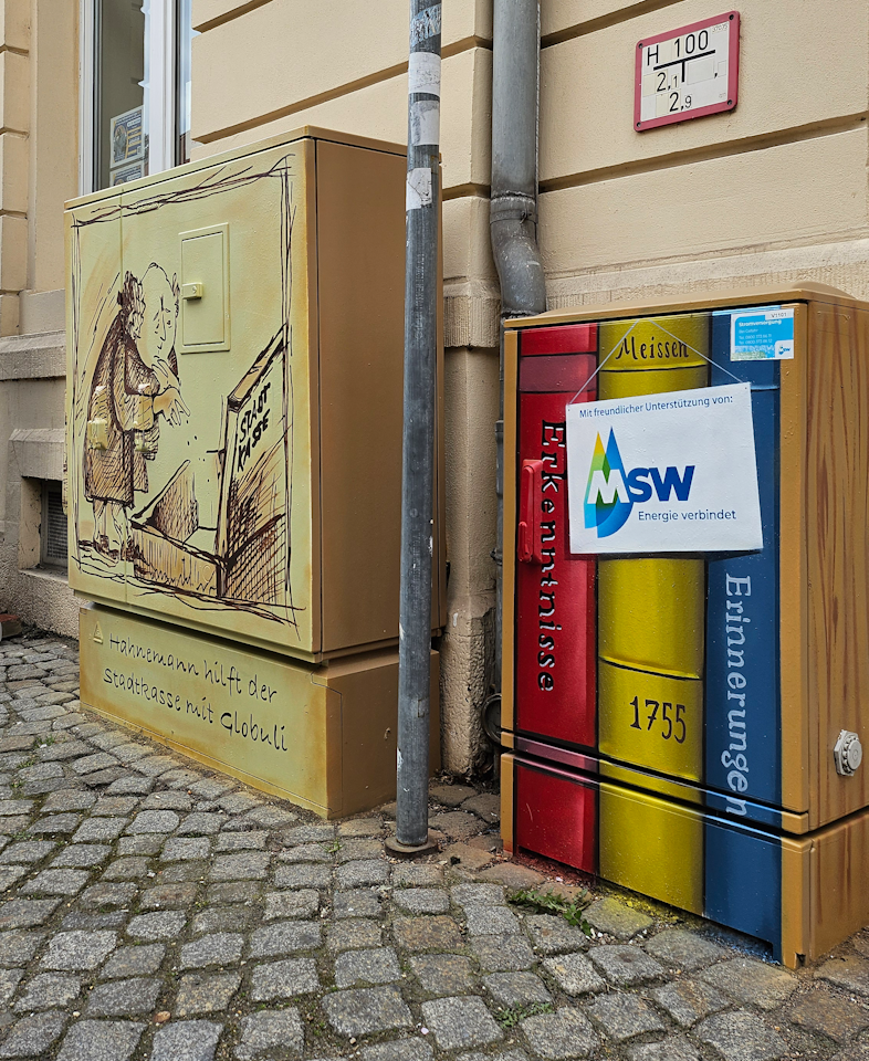 Zwei bemalte Stromkästen stehen auf einem Pflastersteinweg neben einem Gebäude, wobei der linke Kasten ein antikes Motiv mit einer Zeichnung eines Mannes zeigt und der rechte Kasten in einem bunten Buchrücken-Design mit einer MSW-Werbung gestaltet ist.