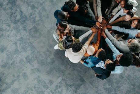 Eine vielfältige Gruppe von Menschen steht in einem Kreis und legt die Hände übereinander in einer unterstützenden Geste der Teamarbeit und Einheit auf einem grauen Teppichboden.