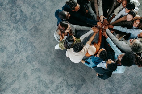 Eine vielfältige Gruppe von Menschen steht in einem Kreis und legt die Hände übereinander in einer unterstützenden Geste der Teamarbeit und Einheit auf einem grauen Teppichboden.