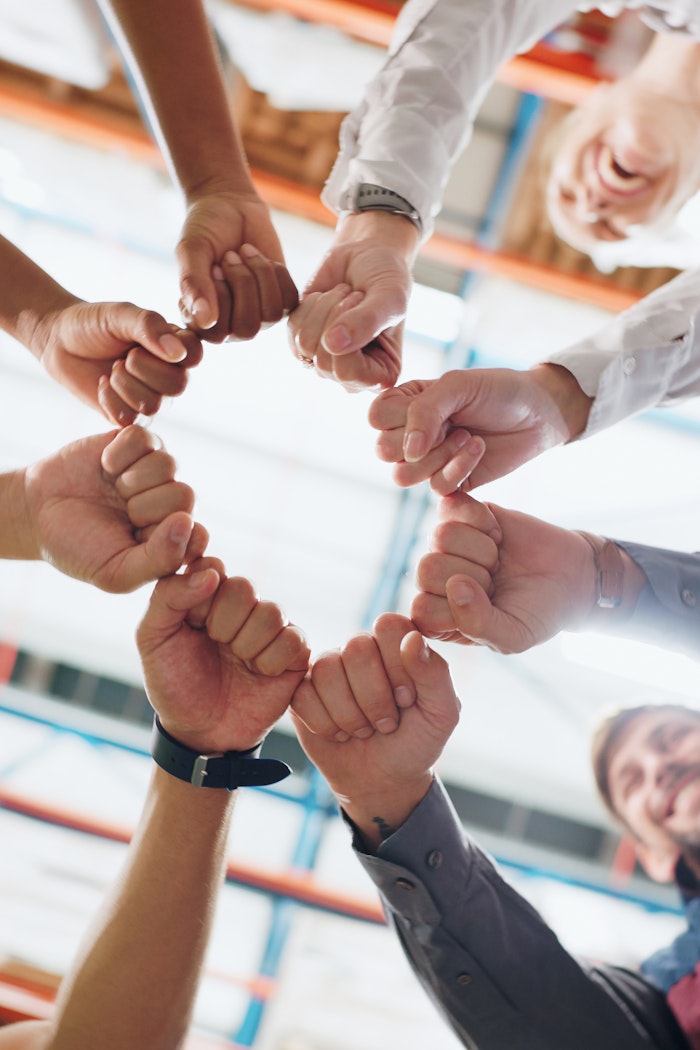 Mehrere Menschen strecken ihre Arme nach vorne und berühren sich mit den Fäusten in einem Kreis, was Zusammenhalt und Teamarbeit symbolisiert.