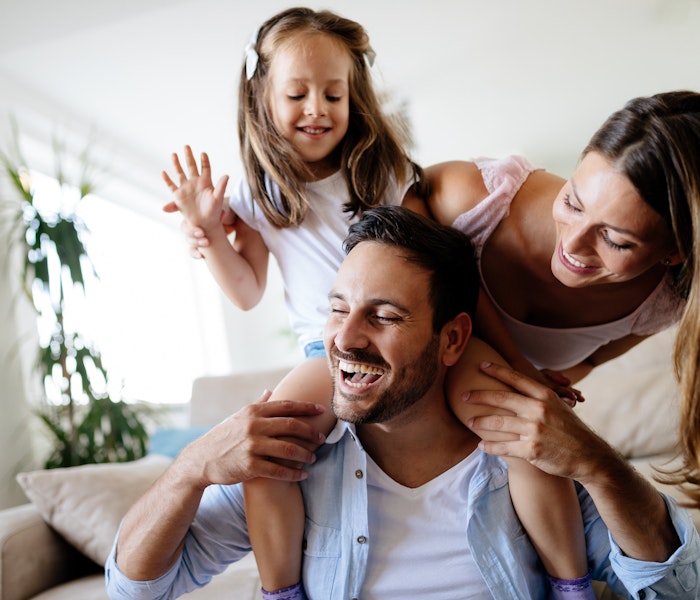 Eine fröhliche Familie spielt zusammen im Wohnzimmer, während das kleine Mädchen auf den Schultern des Mannes sitzt und alle lachen; im Hintergrund steht eine große Zimmerpflanze.
