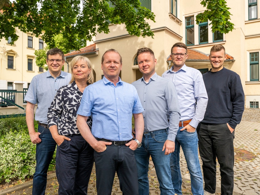 Eine Gruppe von sechs Personen steht lächelnd vor einem Gebäude im Freien, umgeben von grünen Bäumen und Pflastersteinen, während sie legere Kleidung tragen.