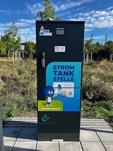 Ein schwarzer Elektroladestation-Schrank steht auf einem gepflasterten Bereich mit grüner Vegetation im Hintergrund, verziert mit Aufklebern des MSW und der Aufschrift "Strom Tank Stelle".
