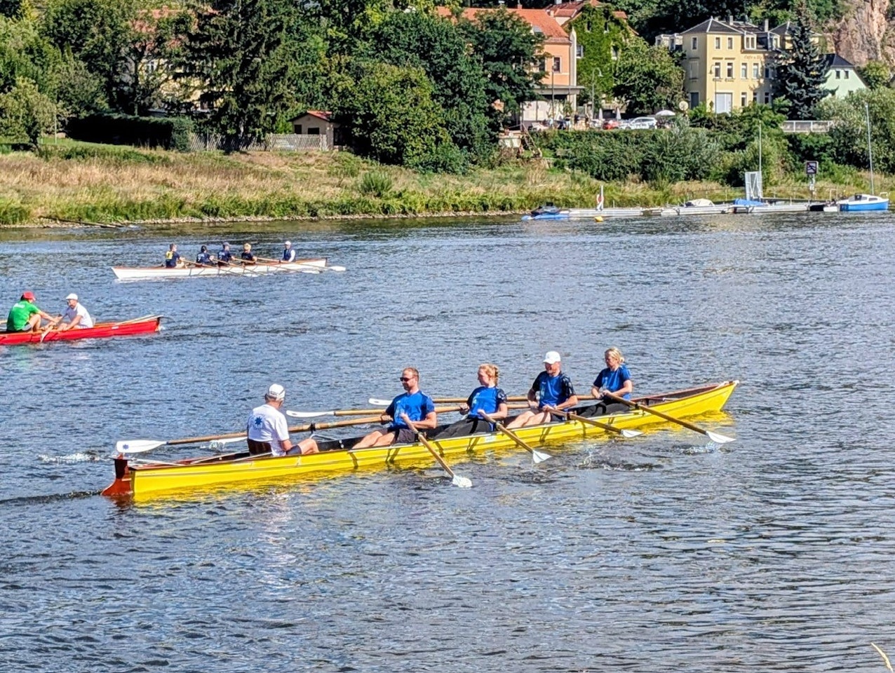 Eine Gruppe von fünf Personen sitzt in einem gelben Ruderboot auf einem ruhigen Fluss, umgeben von üppigem Grün und mehreren Booten im Hintergrund, während sie gemeinsam rudern.
