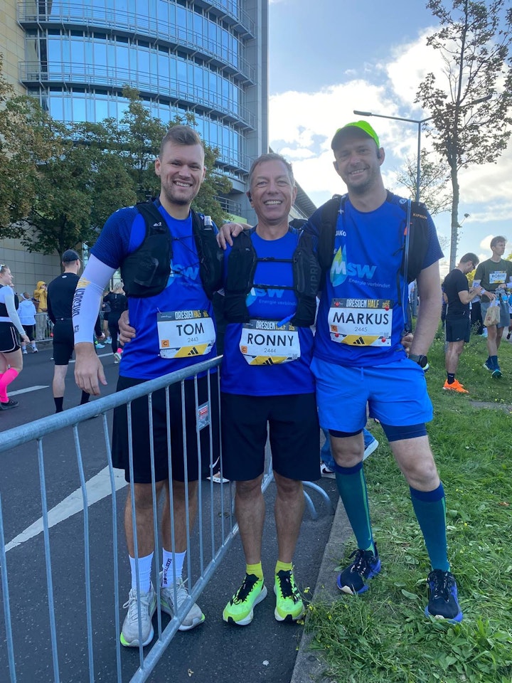Drei Personen in blauen Sportoutfits stehen fröhlich hinter einer Absperrung bei einer Laufveranstaltung auf einer städtischen Straße, während andere Teilnehmer und Grünes im Hintergrund sichtbar sind.