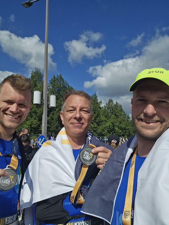 Drei Personen in blauer Sportkleidung lächeln, tragen Medaillen um den Hals und haben weiße Handtücher um die Schultern, während sie unter freiem Himmel bei klarem Himmel posieren; Hintergrund zeigt Bäume und bewölkten Himmel.