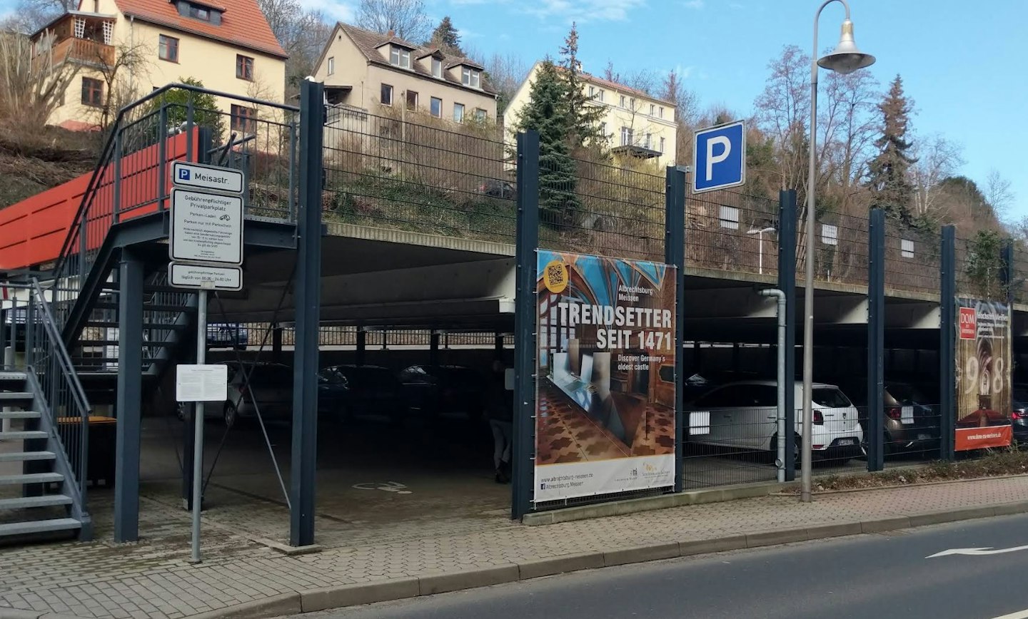 Ein zweistöckiges Parkhaus mit geparkten Autos unter einem blauen P-Schild, umgeben von Wohnhäusern, mit Werbeplakaten und Bäumen im Hintergrund.