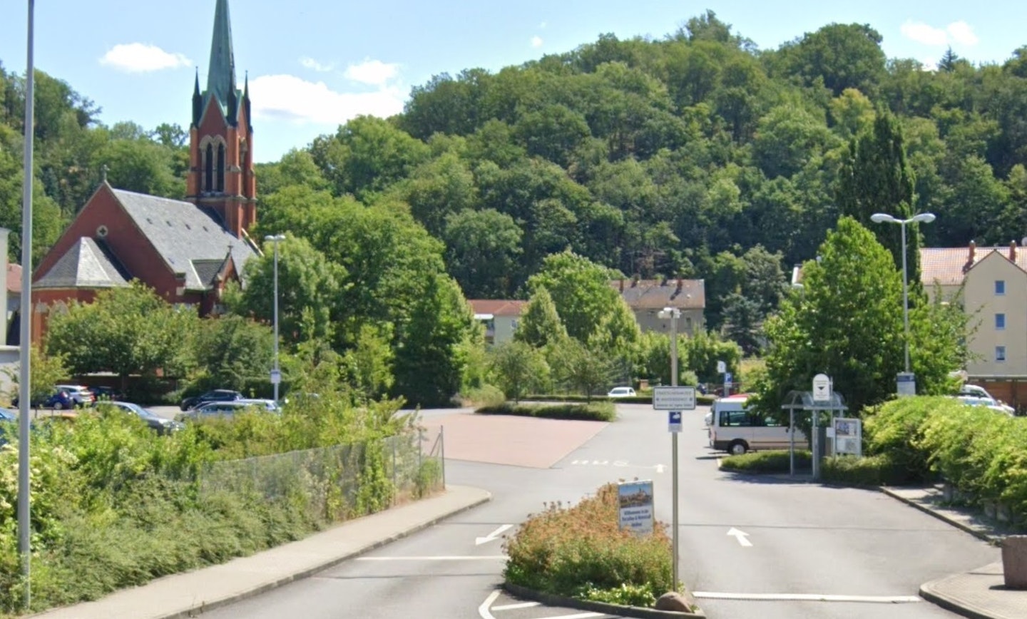 Eine malerische Landschaft mit einem roten Backstein-Gebäude und einem prominenten Kirchturm im Hintergrund, umgeben von üppigen grünen Bäumen und einem Parkplatz im Vordergrund.