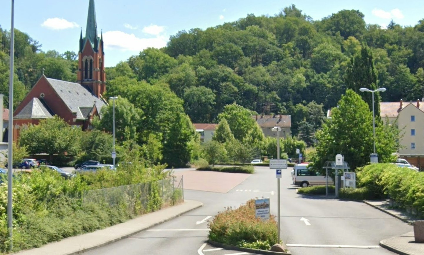 Eine malerische Landschaft mit einem roten Backstein-Gebäude und einem prominenten Kirchturm im Hintergrund, umgeben von üppigen grünen Bäumen und einem Parkplatz im Vordergrund.