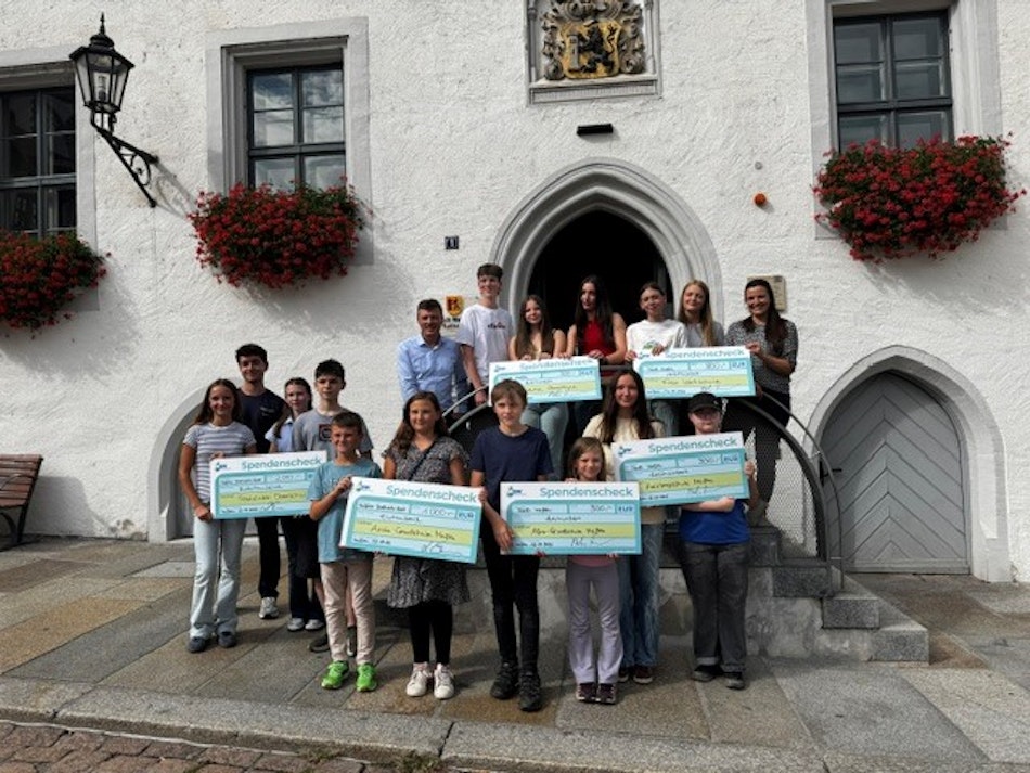In einer malerischen Steinarchitektur vor einem historischen Gebäude mit blumengeschmückten Fenstern posiert eine Gruppe von Menschen, die große Spendenschecks hält, und demonstriert ihre Beteiligung an einer wohltätigen Veranstaltung.