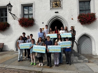 In einer malerischen Steinarchitektur vor einem historischen Gebäude mit blumengeschmückten Fenstern posiert eine Gruppe von Menschen, die große Spendenschecks hält, und demonstriert ihre Beteiligung an einer wohltätigen Veranstaltung.