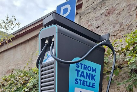 Eine Ladestation für Elektrofahrzeuge steht vor einer mit Efeu bewachsenen Mauer mit einem Schild "Strom Tank Stelle" und einem blauen "P"-Parkplatzzeichen.