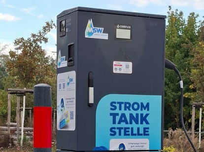Eine schwarze Elektroladestation mit einem Display und einem Ladekabel zeigt Aufkleber der Marke "MSW" und befindet sich im Freien vor Bäumen.