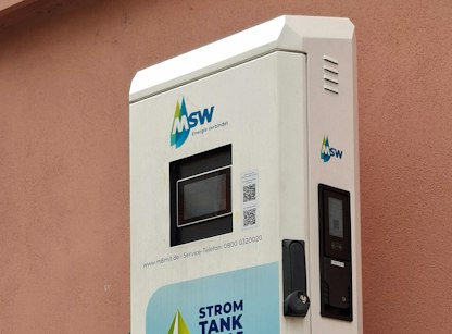 Eine weiße Stromladesäule mit MSW-Branding steht vor einer rosa Wand, ausgestattet mit einem digitalen Display und einem QR-Code auf der Vorderseite.