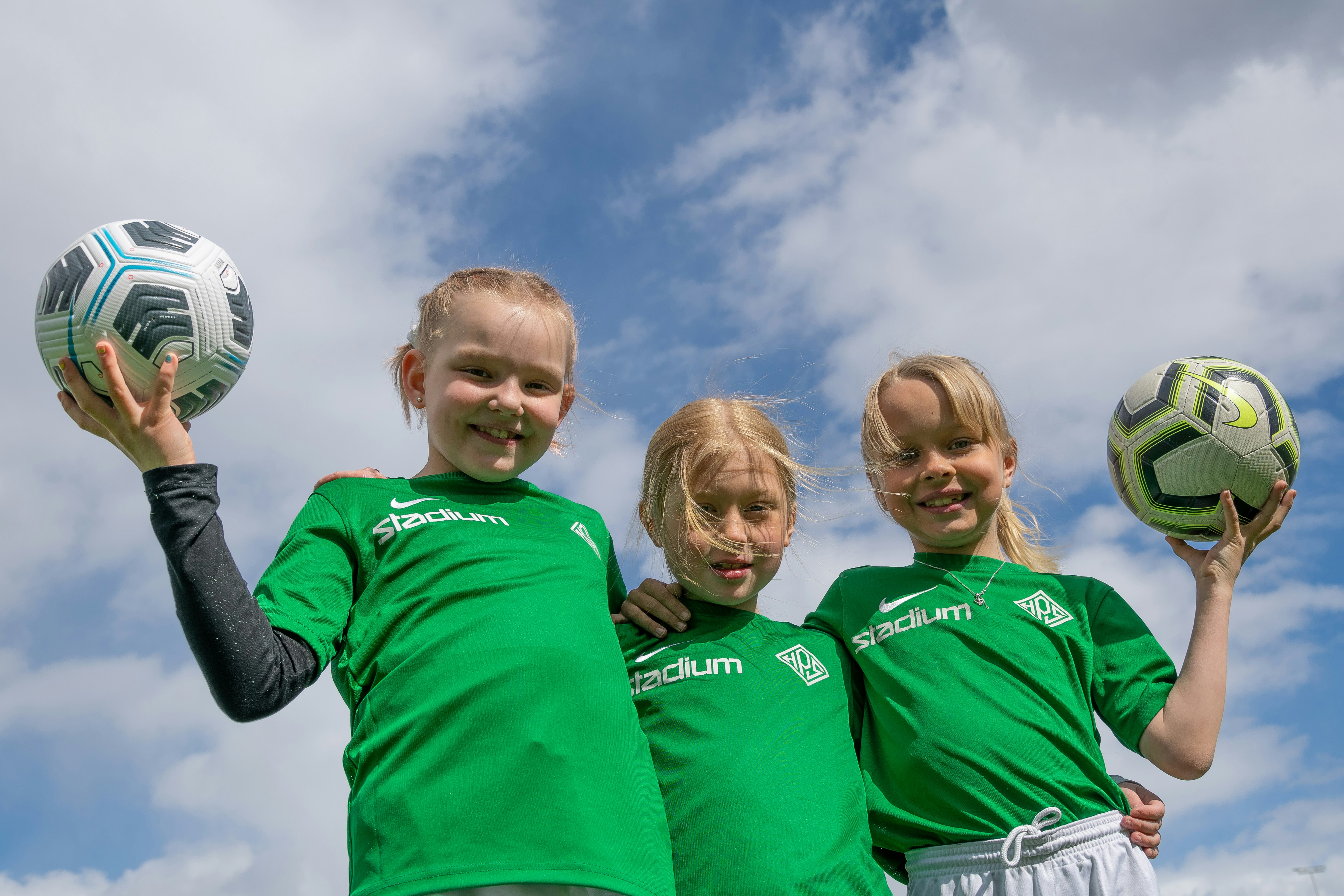 Kolme tyttöä vihreissä pelipaidoissa. Kahdella tytöistä on kädessä jalkapallo. 