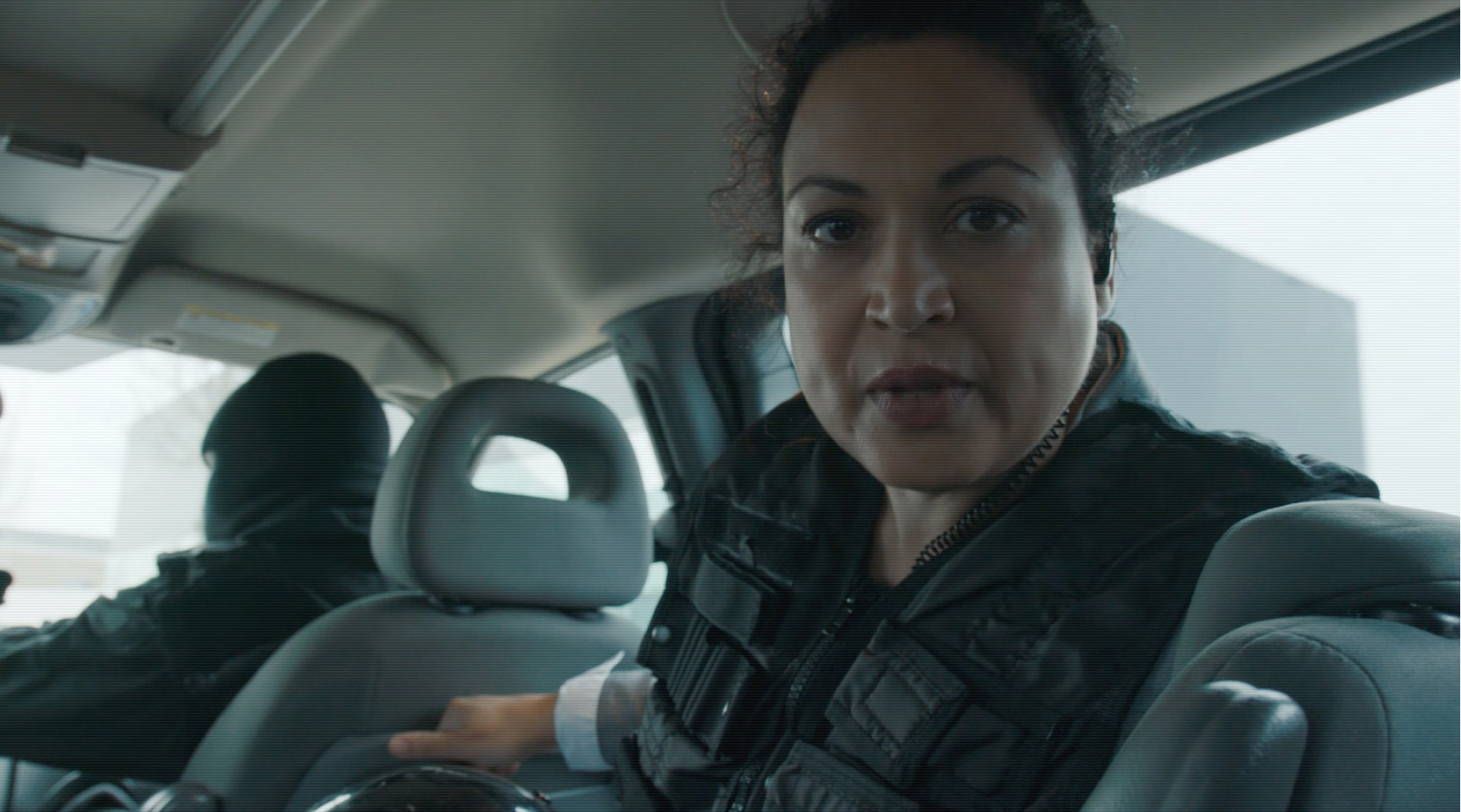 Screencapture of a woman in the back of a car looking into the camera