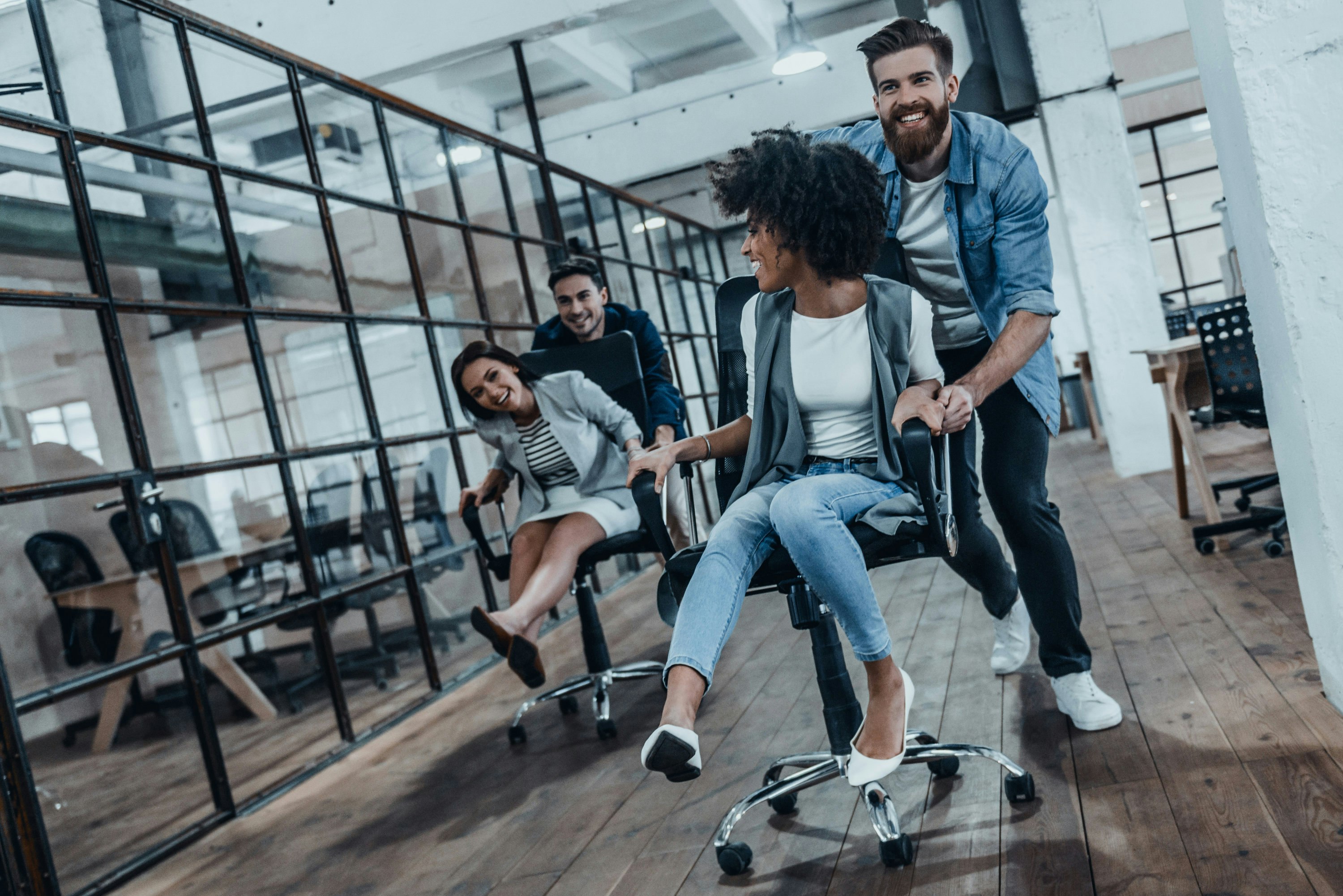 Unstoppable team! Four young cheerful business people in smart casual wear having fun while racing on office chairs and smiling.