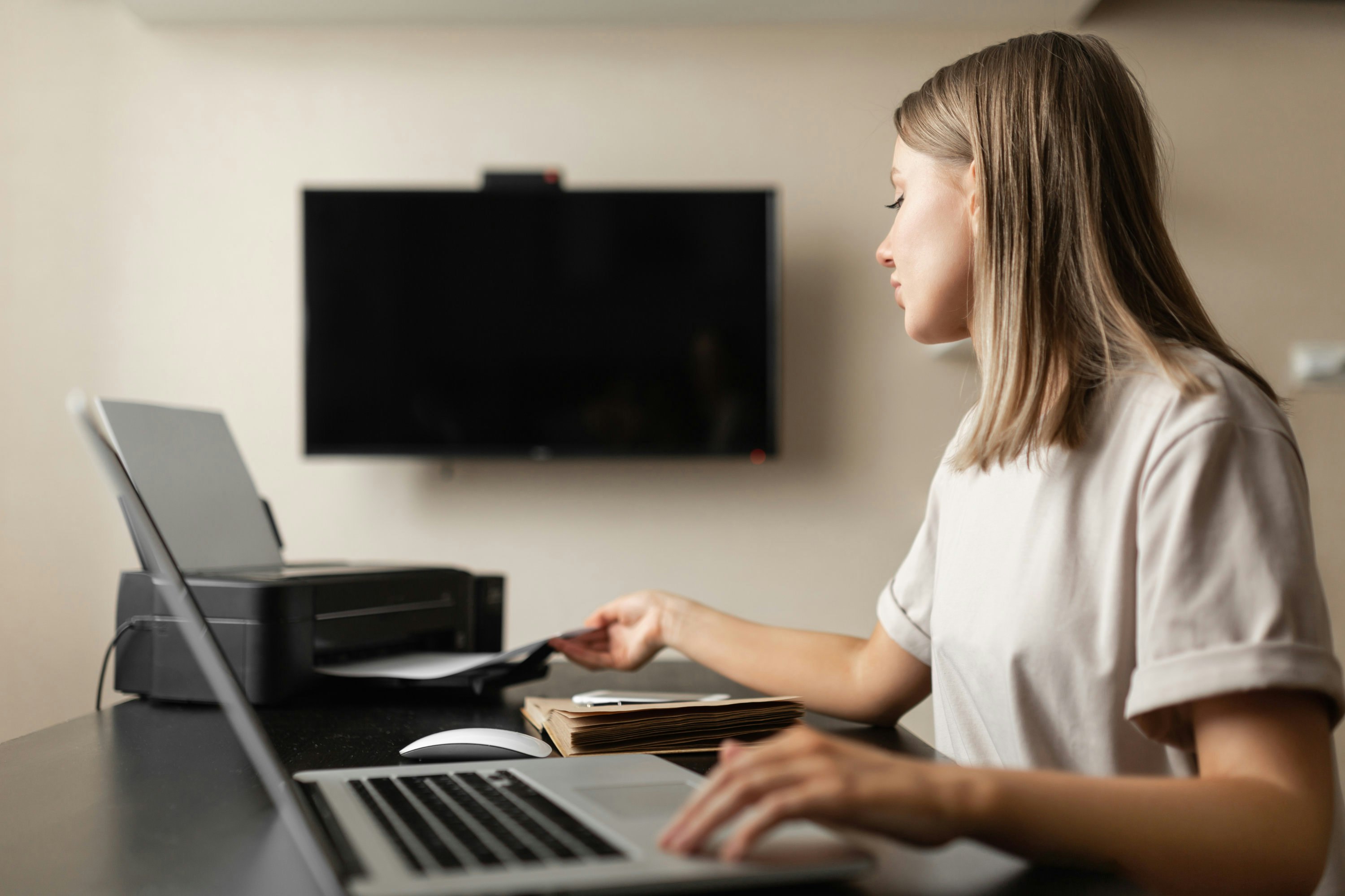 Eine Frau sitzt am Schreibtisch, arbeitet am Notebook und druckt Unterlagen aus. Im Hintergrund ist ein TV an der Wand zu sehen.