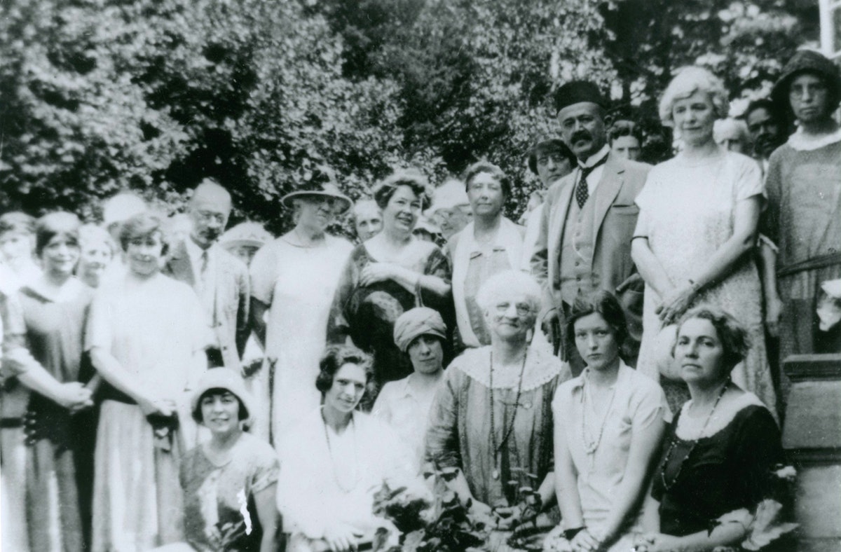 Baha'is who were later named Hands of the Cause of God are pictured at Eliot, Maine, United States, circa 1926, when Marion Jack (standing center in dark gown) was present. Those later elevated to the rank of Hand of the Cause include Mary Maxwell (later Madame Ruhiyyih Rabbani), seated second from the right; Paul Haney (partially obscured in back row to the right of man in eastern-style hat); Louis Gregory, standing second from right next to his wife, Louisa, who was the first Baha'i pioneer to Bulgaria. May Maxwell, a prominent Baha’i and mother of Mary Maxwell, stands at the left in dark gown with hands together. Physician and educator Dr. Susan Moody is seated to the left of Mary Maxwell. Stanwood Cobb, a Baha'i author, is wearing a suit and is standing near the left of the group.