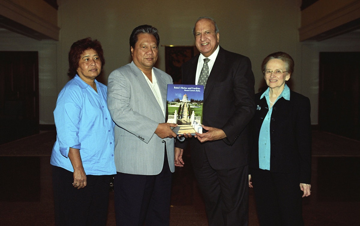A member of the Universal House of Justice, Dr. Peter Khan, second from right, presenting President Kessai Note of the Marshall Islands with a book illustrating the Shrine of the Bab and its surrounding terraces. At left is Mrs. Mary Note and, at right, Dr. Janet Khan.