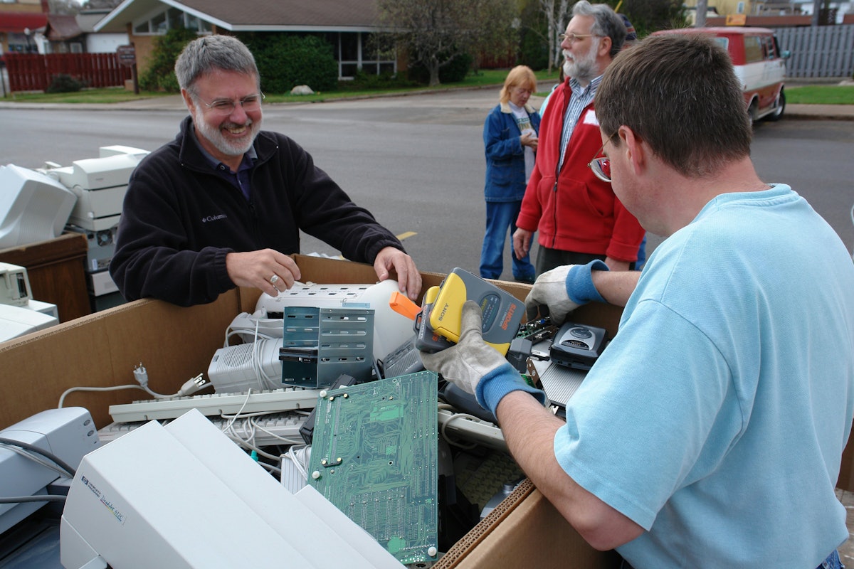 Dr. Rodney Clarken, a Baha'i and one of the original signers of the Earth Keeper Covenant, at left, looks on as "e-waste" is collected in a recycling bin in Marquette, Michigan. Also shown are Jean Soderberg (background, center) and Dennis McCowen (in red). All are Baha'is and all were participating in the Earth Keeper Clean Sweep on 22 April 2006. (Photo by Greg Peterson.)