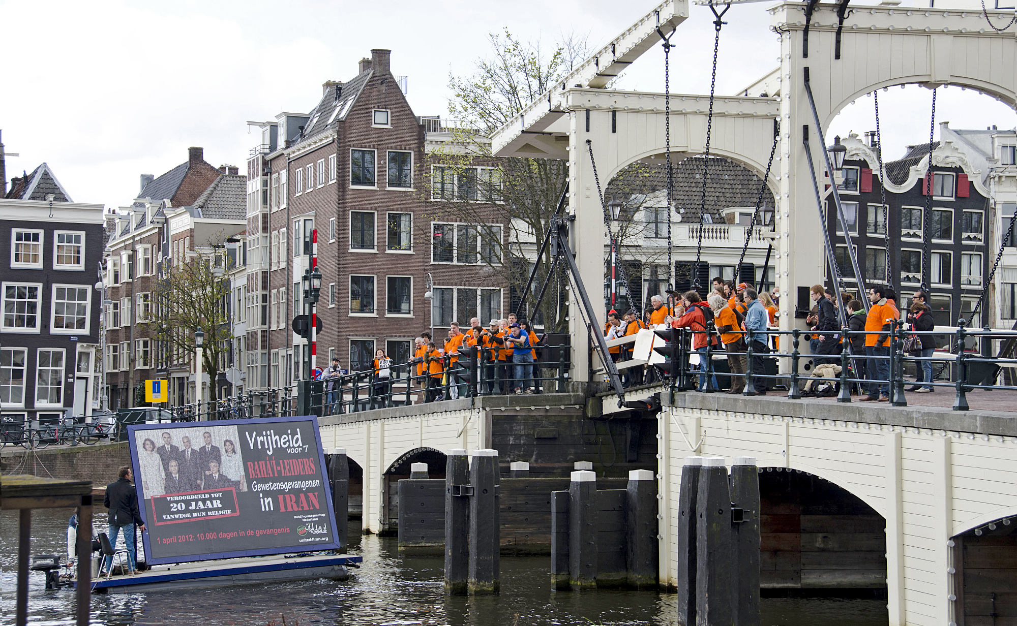 یک تابلوی اعلانات با تصویر هفت رهبر بهائی ایران هنگام عبور از زیر پل ماجره براگ آمستردام، در حالی که حامیان با جلیقه‌های با چاپ نارنجی بر تن، آن را مینگریستند.