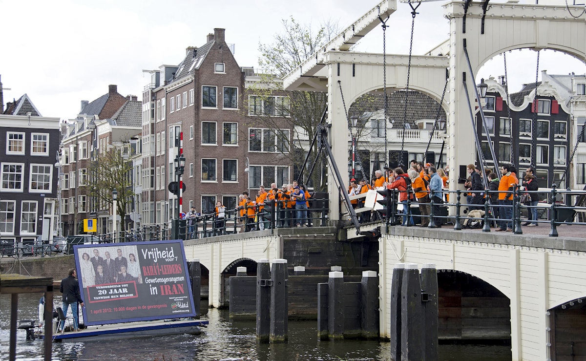 In the Netherlands, a billboard depicting Iran's seven Baha'i leaders passes under Amsterdam's Magere Brug watched by supporters in printed orange vests.