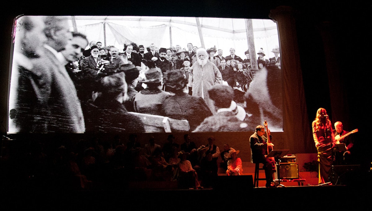 A photograph of 'Abdu'l-Baha speaking at the cornerstone ceremony for the Baha'i House of Worship, Wilmette, is projected at the event's centenary commemoration, held in Chicago on 28 April 2012. Grammy-nominated singer Tierney Sutton is also seen performing with guitarist Jamie Findlay and bassist Kevin Axt.