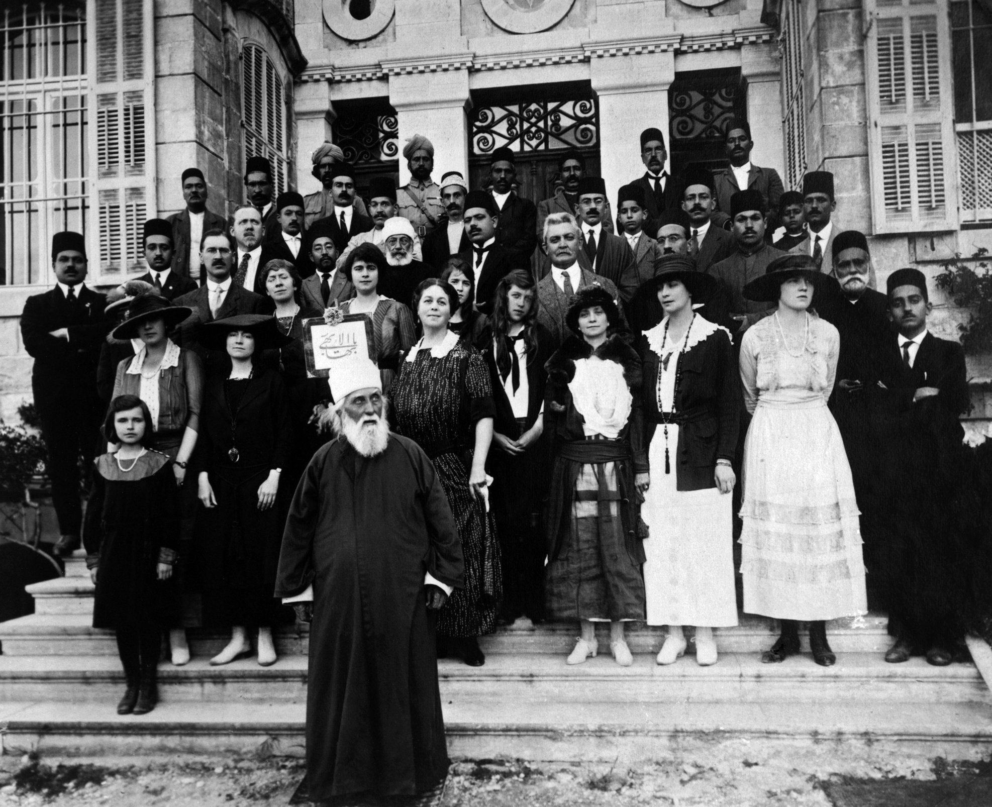 A group of Baha’is and others stand with ‘Abdu’l-Baha outside His house in Haifa. Two Indian soldiers can be seen in the back row. (circa 1918)