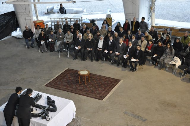 Members of institutions and agencies of the Baha'i community look on as a silver container holding dust from the Shrine of the Bab is placed in the wood-carved rendering of the Greatest Name.