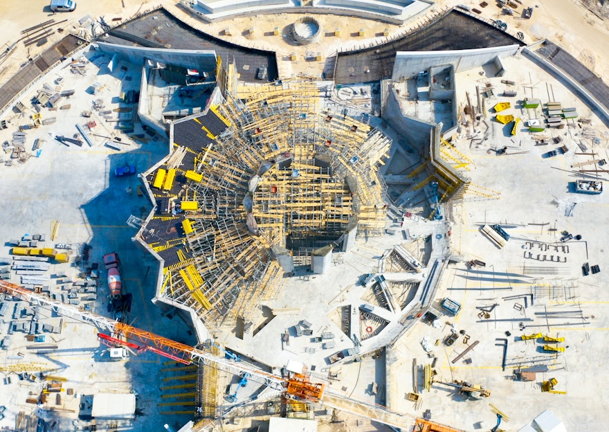 Les huit piliers de l’édifice principal sont maintenant terminés et les échafaudages sont en cours d’installation dans l’espace entre les piliers et les murs plissés achevés, pour permettre les travaux sur le treillis qui surplombera la place centrale.