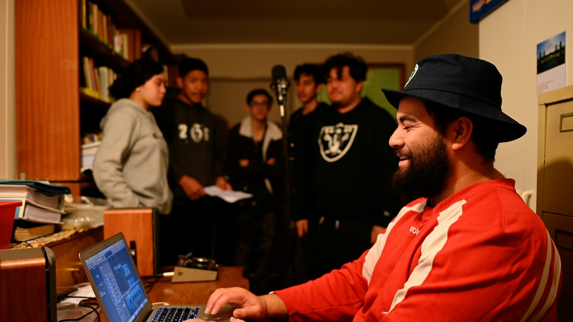 Photo of a recording session in a home studio in Manurewa. A young man in red sits at a computer in the foreground. A group of youth stand around a microphone in the background.