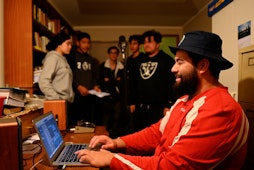 Photo of a recording session in a home studio in Manurewa. A young man in red sits at a computer in the foreground. A group of youth stand around a microphone in the background.