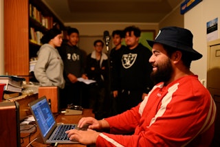Photo of a recording session in a home studio in Manurewa. A young man in red sits at a computer in the foreground. A group of youth stand around a microphone in the background.